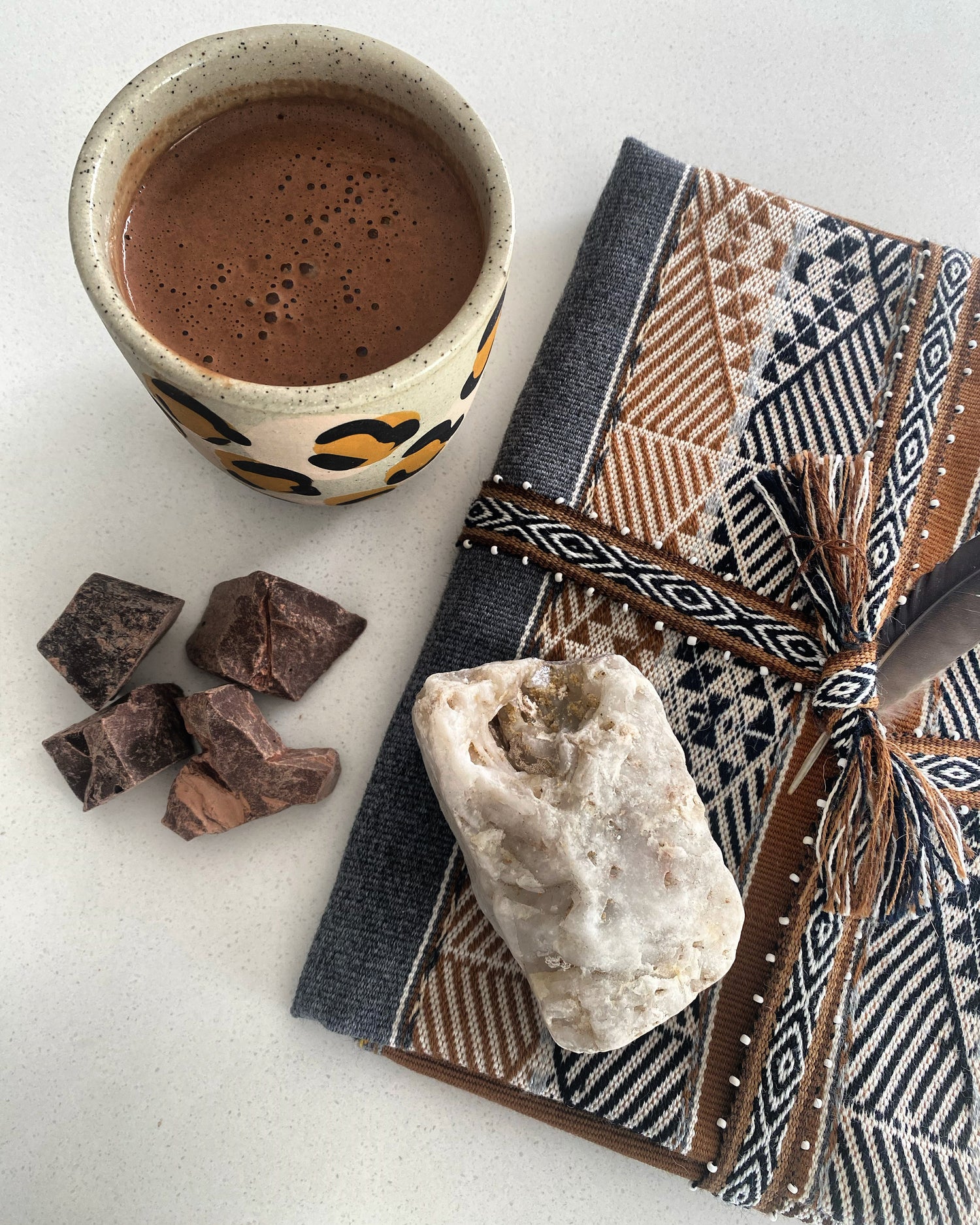 Cup of hot Cacao with cacao pieces and a sacred mesa bundle on a white surface, topped with some natural quartz.