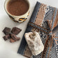 Cup of hot Cacao with cacao pieces and a sacred mesa bundle on a white surface, topped with some natural quartz.