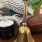Brass bell on a wooden surface with peacock feathers, resin and a dried cacao pod in the background.
