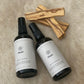Two bottles laying flat of 'Pavana' mist with three raw sticks of Palo Santo on a beige cow hide.