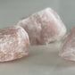 Three rose quartz chunks laying on a stone surface, light filtering from the right and showing a side view.
