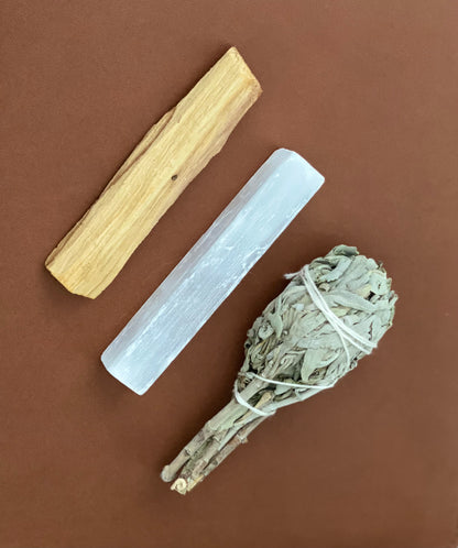 A smudge kit consisting of a white sage stick, a palo santo stick, and a selenite crystal stick, all arranged on a brown flat surface.