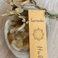 'Surrender'  Incense Packaged laying on top of a bowl of tumbled citrine crystals and three dried flowers.