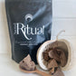 A 300g black package of Sacred Ritual brand Ceremonial Cacao with small chunks of cacao displayed in front of it, all placed on a white surface with a decorative pod to the side.