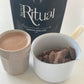 A latte mug of cacao with a small pouring pot of raw Cacao pieces with a bag of Ceremonial Cacao labelled 'Sacred Ritual' in the background.