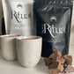A 1kg bag of ceremonial grade cacao next to two cups of prepared cacao and loose cacao chunks on a light-colored surface.