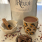 Ceremonial grade cacao in a leopard print mug, bowl of cacao pieces, and sticks of Palo Santo on a light surface.