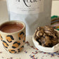 A cup of hot Cacao in a leopard print mug, next to a small bowl with chumpi stones, on a surface scattered with flower petals, and a  1kg bag of Ceremonial Cacao packaged and  labeled ' Sacred Ritual' in the background.