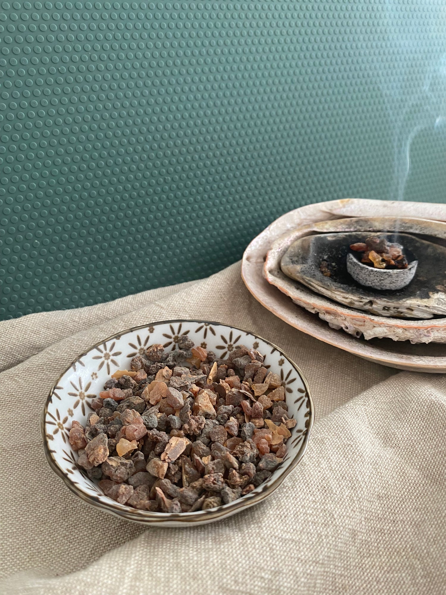 Two ceramic containers with resin, one is a bowl, the other is a shell with burning myrrh on a charcoal disc and smoke with  a textured green background. 