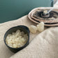White Copal burning on a charcoal disc in the background showing a small black tin of white copal pieces in front, close up on beige linen surface.