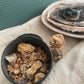 Small black tin of dark copal, with deep earthy tones from sand to Cola in the foreground and some burning resin on a charcoal disc blurred in the background.