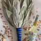 Dried leaf rattle tied with a vibrant Peruvian ribbon on a white surface with dried flowers scattered around