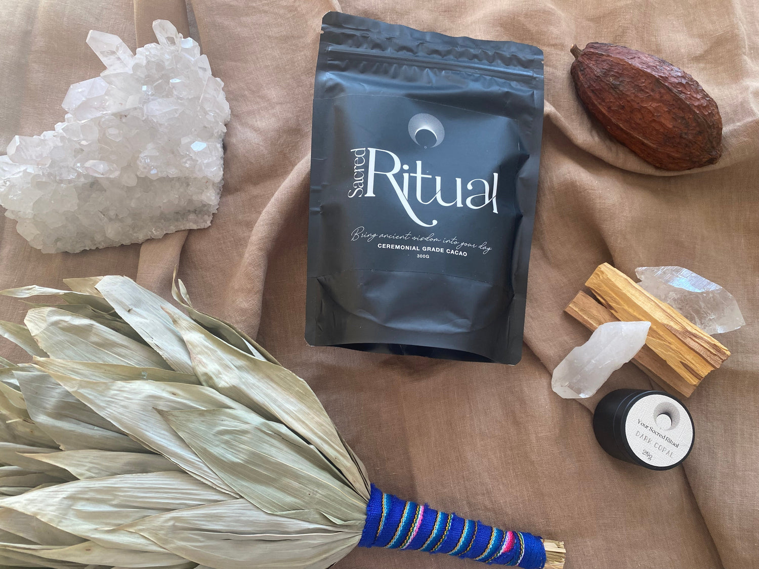 300g bag of ceremonial cacao in a black pouch surrounded by ceremonial tools including cacao pod, quartz cluster, and points, a leaf rattle and some palo santo and dark copal resin. All laid out beautifully on a natural fabric.